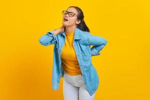 Beautiful young Asian woman student in denim clothes suffering from backache isolated on yellow background. People sincere emotions lifestyle concept