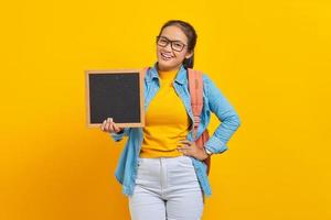 Portrait of smiling young Asian woman student in casual clothes with backpack holding blank chalkboard isolated on yellow background. Education in college university concept