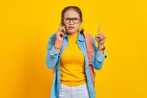 Portrait of annoyed young Asian woman student in casual clothes with backpack talking on mobile phone and pointing finger up isolated on yellow background. Education in college university concept photo