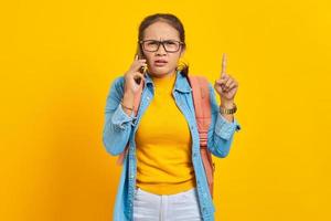 retrato de una joven estudiante asiática molesta con ropa informal con mochila hablando por teléfono móvil y señalando con el dedo aislado en un fondo amarillo. educación en concepto de universidad universitaria foto