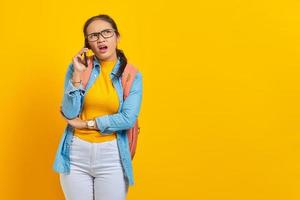 Portrait of confused young Asian woman student in casual clothes with backpack talking on smartphone and looking at copy space isolated on yellow background. Education in college university concept photo