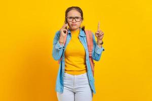 Portrait of confused young Asian woman student in casual clothes with backpack talking on mobile phone and pointing finger up isolated on yellow background. Education in college university concept photo