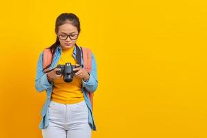retrato de una joven asiática pensativa vestida de denim y mirando una foto en una cámara aislada de fondo amarillo. pasajero que viaja los fines de semana. concepto de viaje de vuelo aéreo