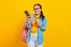 Portrait of smiling young Asian woman student in denim clothes with backpack holding cup of coffee and using smartphone isolated on yellow background photo
