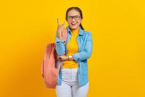 hermosa joven estudiante asiática vestida de denim con mochila gritando con expresión loca haciendo símbolo de rock con las manos arriba aisladas en fondo amarillo. educación en concepto de colegio universitario foto