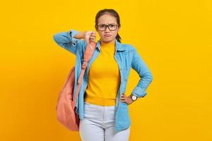 Annoyed young Asian student in denim outfit with backpack showing thumbs down with finger isolated on yellow background. Education in high school university college concept photo