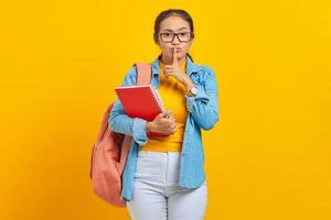 Beautiful young woman student in denim clothes with backpack and holding book, fingers covering lips, making silent gesture on yellow background. Education in high school university college concept photo