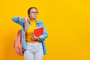 Sad young Asian woman student in denim clothes with backpack holding notebook, showing thumb down while looking aside isolated on yellow background. Education in high school university college concept photo