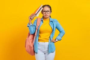 Pensive young Asian woman student in denim clothes with backpack holding notebook isolated on yellow background. Education in high school university college concept photo