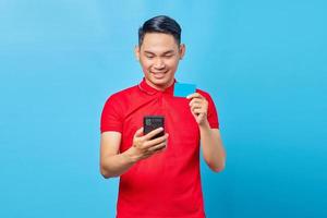 Portrait of smiling asian young man using mobile phone and showing credit card isolated blue background photo