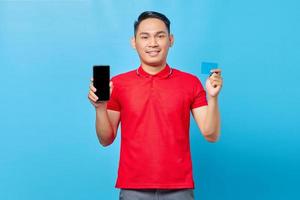 retrato de un joven asiático sonriente que muestra un teléfono móvil con pantalla en blanco y una tarjeta de crédito aislada de fondo azul foto