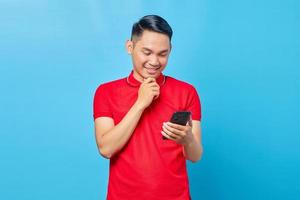 Portrait of smiling Asian young man looking at mobile phone and holding chin isolated on blue background photo