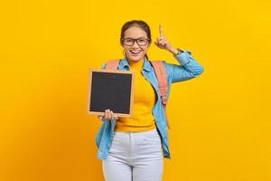 Portrait of smiling young Asian woman student in casual clothes with backpack holding blank chalkboard and pointing up with finger isolated on yellow background photo
