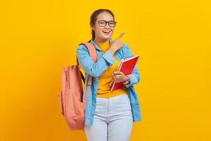 retrato de una joven estudiante asiática sonriente con ropa informal con mochila sosteniendo un libro y señalando a un lado con el dedo aislado en un fondo amarillo. educación en concepto de universidad universitaria foto