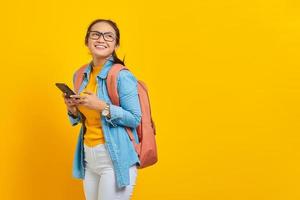 Portrait of smiling young Asian woman student in casual clothes with backpack using mobile phone and looking aside isolated on yellow background. Education in college university concept photo