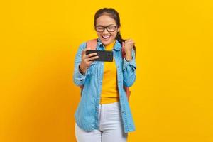 retrato de una alegre joven estudiante asiática vestida de forma informal con mochila jugando videojuegos en el teléfono móvil y celebrando el éxito aislada de fondo amarillo foto