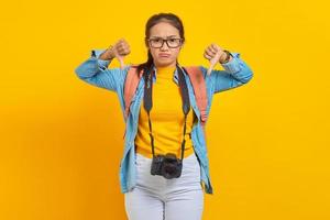 Portrait of angry young traveler Asian woman with backpack and camera in denim clothes while showing thumb down with fingers isolated on yellow background. Air flight journey concept photo