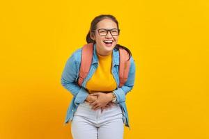 hermosa joven estudiante asiática vestida de denim con mochila mostrando una fuerte risa con la mano en el estómago aislada en un fondo amarillo. educación en concepto universitario foto