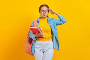 hermosa joven estudiante vestida de denim con mochila, sosteniendo un cuaderno y mostrando un gesto de respeto con la mano aislada en el fondo amarillo. educación en el concepto de colegio universitario de secundaria