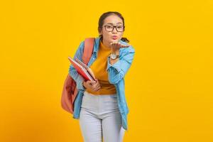 Beautiful young woman student in denim clothes with backpack and holding book, hold hand palms send air kiss to boyfriend on yellow background. Education in high school university college concept photo