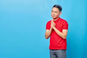 Portrait of handsome Asian young man wearing red shirt praying with hands together asking for forgiveness isolated on blue background photo