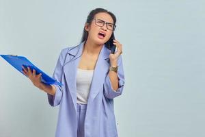 Portrait of surprised young Asian woman talking on mobile and looking at task list in documents folder isolated on purple background photo