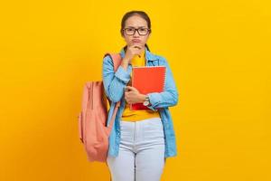 Portrait of pensive young Asian woman student in casual clothes with backpack holding book and thinking about question isolated on yellow background. Education in college university concept photo