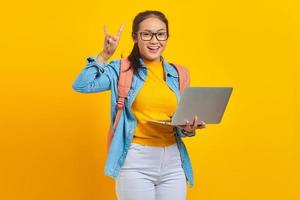 Portrait of cheerful young Asian woman student in casual clothes with backpack using laptop and doing rock symbol with hands up isolated on yellow background. Education in university college concept photo