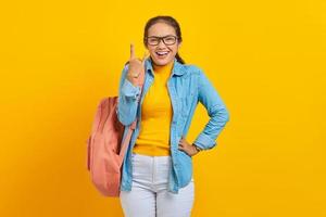 hermosa joven estudiante asiática vestida de denim con mochila gritando con expresión loca haciendo símbolo de rock con las manos arriba aisladas en fondo amarillo. educación en concepto de colegio universitario foto