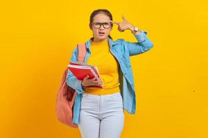 Beautiful young woman student in denim clothes with backpack,holding note book and Point fingers to head as if she about shoot herself isolated on yellow background photo