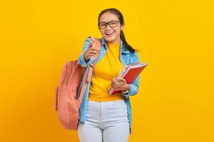 Portrait of excited young Asian woman student in denim clothes with backpack holding notebook, pointing fingers at camera on yellow background. Education in high school university college concept