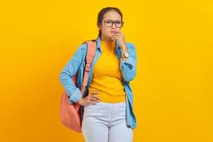 Beautiful young Asian student in denim outfit with backpack and looking serious thinking about question isolated on yellow background. Education in high school university college concept photo