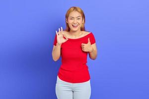 Portrait of beautiful cheerful Asian woman wearing red dress smiling while showing ok sign and gesture of approval with finger on purple background photo