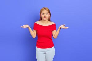 Portrait of attractive confused young Asian woman with short blonde hair in red dress standing and shrugging shoulders isolated over purple background photo