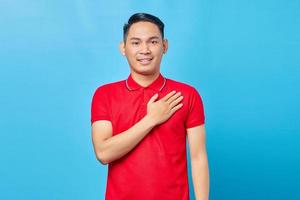 retrato de un apuesto joven asiático con camisa roja tomándose de la mano en el pecho para agradecer a alguien y expresar gratitud aislado de fondo azul