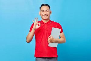 Portrait of smiling Asian young man in red shirt holding laptop and making okay gesture, showing approval symbol isolated on blue background photo