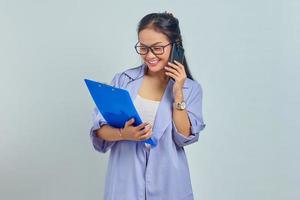 Portrait of cheerful young Asian woman laughing while talking on mobile phone with friends about something funny while holding document folder isolated on purple background photo