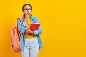 retrato de una joven estudiante asiática pensativa vestida de forma informal con mochila sosteniendo un libro y pensando en una pregunta aislada en un fondo amarillo. educación en concepto de universidad universitaria foto