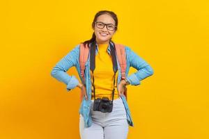 Portrait of cheerful young traveler Asian woman with backpack and camera in denim clothes isolated on yellow background. Passenger traveling on weekends. Air flight journey concept