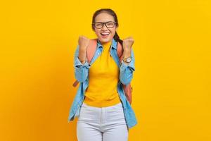 Excited young Asian woman student in denim outfit with backpack standing doing winning gesture celebrating isolated on yellow background. Education in university concept photo