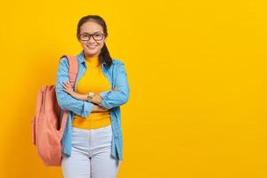 joven estudiante asiático sonriente con traje de mezclilla con mochila cruzada en el pecho y con aspecto confiado aislado en un fondo amarillo. educación en el concepto de colegio universitario de secundaria foto
