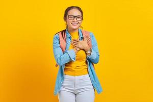 sonriente joven estudiante asiática vestida de denim con mochila con las manos cruzadas en el pecho y el corazón aislado en un fondo amarillo. educación en concepto universitario foto