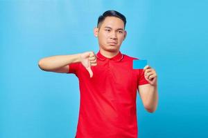 retrato de un joven asiático sonriente que sostiene una tarjeta de crédito y muestra un gesto con el pulgar hacia abajo sobre fondo azul foto