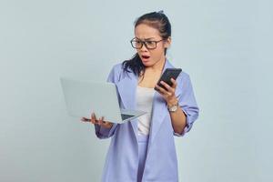 Portrait of surprised young Asian woman holding smartphone and looking at incoming email on laptop isolated on white background photo