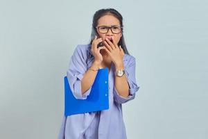 Portrait of cheerful young Asian woman laughing while talking on mobile phone with friends about something funny while holding document folder isolated on purple background photo