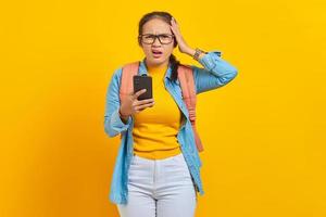 Portrait of surprised young asian woman student in casual clothes  with backpack using mobile phone with open mouth and holding hands in head isolated on yellow background photo