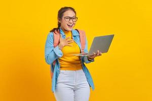 Portrait of excited young Asian woman student in casual clothes with backpack using laptop with hand on chest isolated on yellow background. Education in university college concept photo