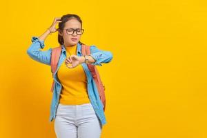 confundida joven estudiante asiática vestida de denim con mochila revisando el tiempo de guardia aislada de fondo amarillo. educación en concepto de colegio universitario foto