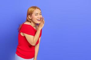 retrato de una joven asiática vestida de rojo mirando la cámara y susurrando mientras sostenía la mano en la boca aislada sobre un fondo morado foto