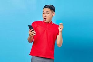 Portrait of surprised young asian man holding mobile phone and showing credit card isolated on blue background photo